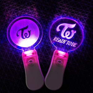 May include: Two purple and pink light-up  TWICE  lightsticks. The lightstick on the right has the text "READY TO BE" on it.