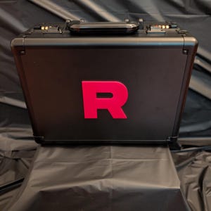 May include: A black briefcase with a red "R" on the front. The briefcase has a handle and metal details. It is on a black surface against a black backdrop.