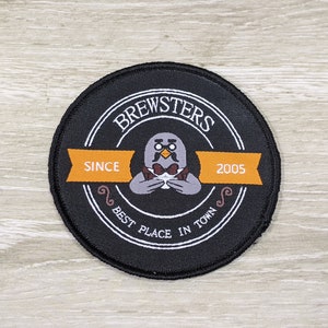 May include: Black and white circular patch with the text "Brewsters" in a white circle with the text "Since 2005" on a yellow banner. The patch also features a cartoon bird wearing a bow tie and holding a cup.