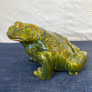 May include: A ceramic toad figurine featuring a glossy finish in shades of deep forest green and olive. The surface is textured with raised bumps and ridges, with subtle hints of reddish-brown coloration around the eyes and along the back.
