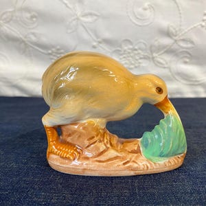May include: A ceramic figurine of a kiwi bird with a light brown body, orange feet, and a long orange beak. The bird is standing on a textured brown base and is pecking at a small, light green, shell-shaped object on the ground.