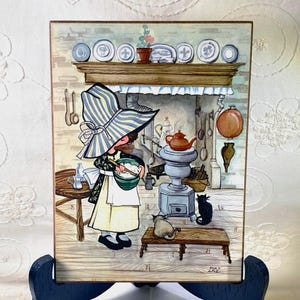 May include: A vintage kitchen scene illustration on a wooden plaque. A figure in a large hat and apron stirs a bowl. A teapot sits atop a stove, with two cats nearby. Decorative plates line a shelf above the fireplace.
