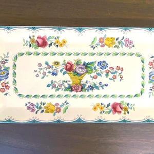 May include: Rectangular serving tray with a floral design. The tray has a cream-colored base with a teal border and scalloped edges. The floral pattern includes a basket of flowers and scattered blooms in pink, yellow, and blue.