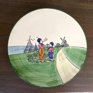 May include: A round decorative plate with a hand-painted scene of two children, a sailboat, and a windmill. The plate has a green border and a cream-colored center. The children are wearing colorful clothing.