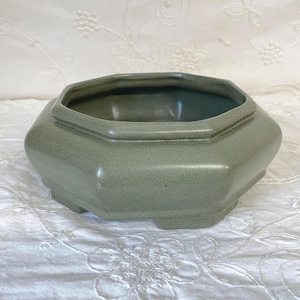 May include: An octagonal ceramic planter with a muted sage green glaze and a smooth, matte finish. The vessel features a wide, open top with a defined rim and a low-profile base with small, integrated feet, presenting a clean and simple form.