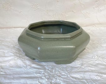 May include: An octagonal ceramic planter with a muted sage green glaze and a smooth, matte finish. The vessel features a wide, open top with a defined rim and a low-profile base with small, integrated feet, presenting a clean and simple form.