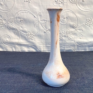 May include: A slender, off-white ceramic vase with a bulbous base and a long, narrow neck. The vase features subtle brown marbling and stands on a dark blue surface. The background includes a white fabric with floral embroidery.