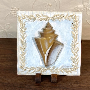 May include: A square, hand-painted artwork featuring a detailed seashell in shades of gold and white against a light blue background. The seashell is framed by a gold-colored laurel wreath design. The artwork is displayed on a wooden easel.