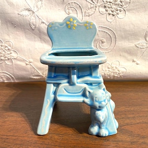 May include: A light blue ceramic chair-shaped planter with a decorative back and a small cat figurine on the front leg. The chair has yellow floral accents and a curved design. The planter is on a wooden surface.