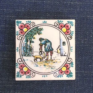 May include: Square ceramic tile with a hand-painted scene of a person, dog, and fish near a fence and building. The tile has a floral border with red, yellow, and green accents. The background is a dark blue fabric.