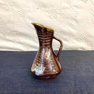 May include: A small, brown ceramic pitcher with a handle and a gold-colored rim. The pitcher has a glossy finish and a textured surface. The background is a white cloth with a floral pattern and a blue surface.