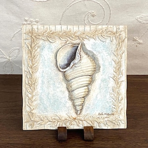 May include: Square, off-white ceramic tile featuring a detailed seashell illustration. The shell is painted in shades of white, beige, and gray, set against a light blue background. The tile is framed with a decorative border of leaves and is displayed on a wooden stand.
