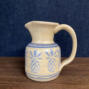 May include: A beige ceramic pitcher featuring a hand-painted blue pineapple pattern around the body. The pitcher has a wide spout, a curved handle on the right side, and horizontal blue bands encircling the top and bottom sections of the vessel.