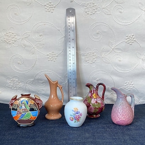 May include: A collection of decorative vases and pitchers in various shapes, sizes, and colors. Includes a brown vase with a unique handle, a light orange vase, a white vase with floral designs, a maroon pitcher with a handle, and a pink pitcher. A ruler is also present.
