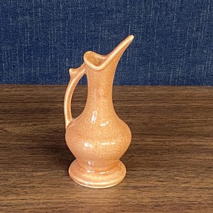 May include: A small, peach-colored ceramic pitcher with a glossy, speckled finish. The vessel features a rounded base, a slender neck, a flared spout, and a single curved handle attached to the side, standing upright on a flat surface.