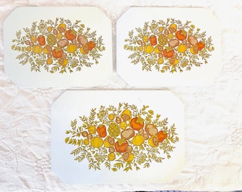 May include: Three white rectangular placemats with clipped corners, each featuring a central botanical illustration of various vegetables, including mushrooms, artichokes, and peppers in shades of orange, yellow, and olive green.