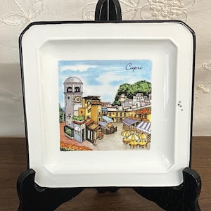 May include: White square decorative plate with a black border. The plate features a colorful illustration of a town square with the word "Capri" written above the image. The image includes buildings, trees, and umbrellas.