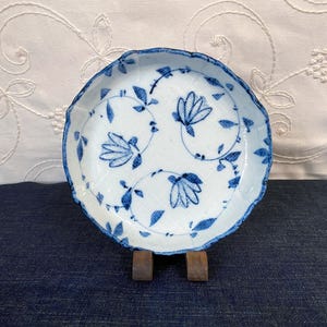 May include: A small, white ceramic plate with a scalloped edge and blue floral designs. The plate features a delicate pattern of stylized flowers and vines. The plate is displayed on a wooden stand against a white background.