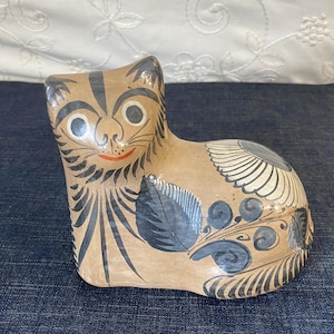 May include: A ceramic cat figurine in a seated position, hand-painted with black and blue details on a tan base. The cat features large eyes, a red smile, and decorative leaf and swirl patterns.