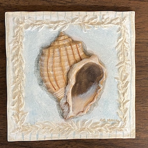 May include: A square, off-white decorative tile featuring a detailed, hand-painted seashell. The shell is a warm tan color with a darker interior, set against a light blue background. The tile is framed with a cream-colored, embossed border. The artist's signature is visible.