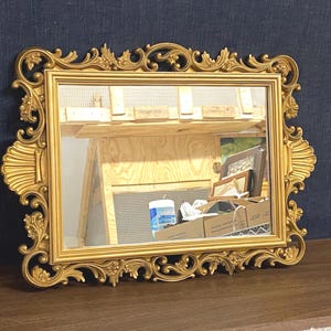 May include: A rectangular mirror featuring an ornate gold-colored frame with intricate scrollwork, floral motifs, and shell-like side accents. The frame surrounds a clear glass reflective surface that shows a reflection of a wooden structure.