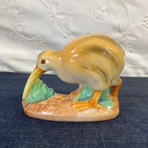 May include: A ceramic figurine of a kiwi bird, featuring a tan body, long beak, and orange feet. The bird is perched on a brown base with green foliage accents. The figurine has a glossy finish.