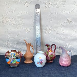May include: Five small ceramic vessels of varying shapes and colors, including a brown piece with a figure, a peach pitcher, a white floral vase, a dark pink floral pitcher, and a textured pink pitcher, next to a 12-inch metal ruler.