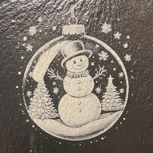 Pode incluir: Enfeite de Natal em ard&oacute;sia preta com um desenho de boneco de neve. O boneco de neve usa um chap&eacute;u alto e um cachecol, rodeado por flocos de neve e pinheiros. O enfeite &eacute; gravado com uma cena de inverno detalhada.