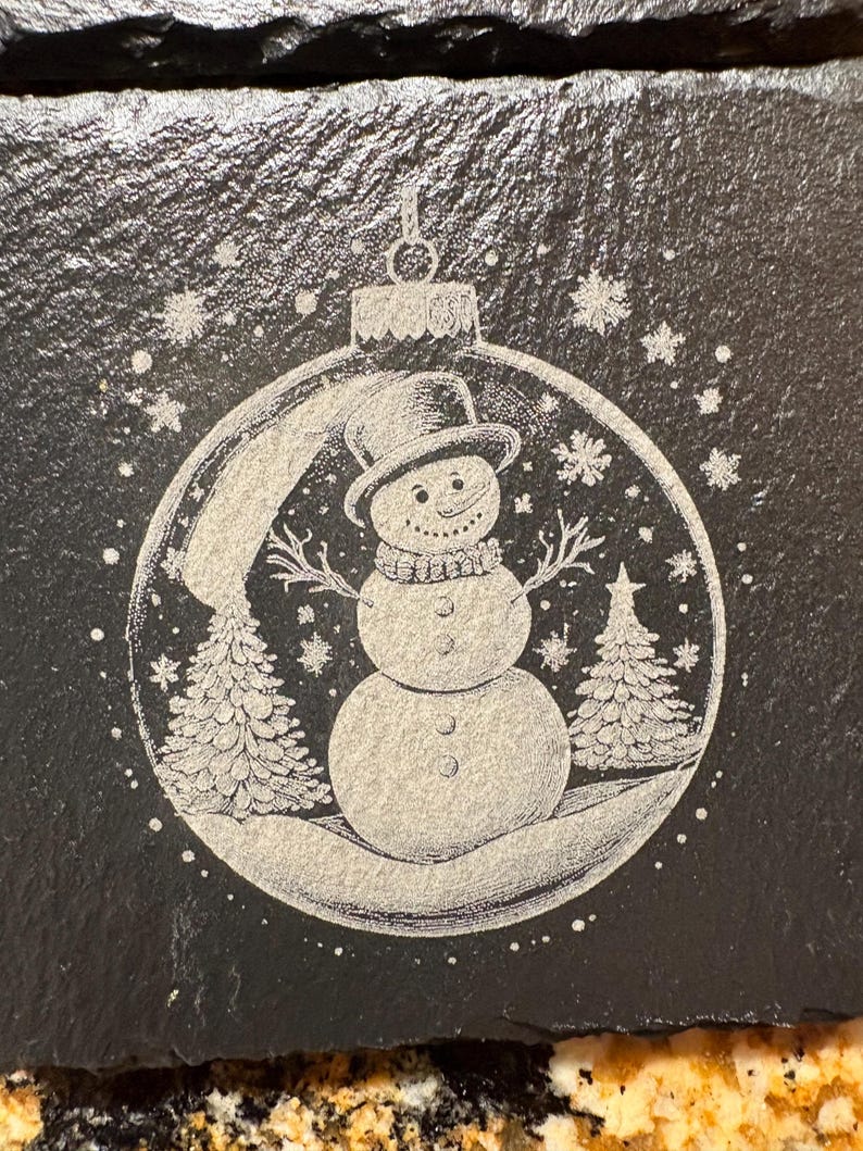 Pode incluir: Enfeite de Natal em ard&oacute;sia preta com um desenho de boneco de neve. O boneco de neve usa um chap&eacute;u alto e um cachecol, rodeado por flocos de neve e pinheiros. O enfeite &eacute; gravado com uma cena de inverno detalhada.