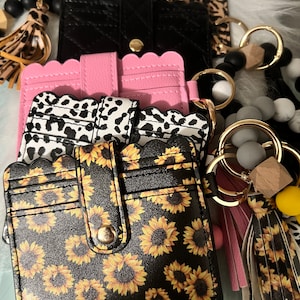 May include: A set of four card holders with different patterns. The card holders are black, pink, black and white leopard print, and black with a sunflower pattern. Each card holder has a gold key ring attached.