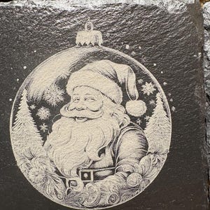 Pode incluir: Conjunto de bases quadradas de ard&oacute;sia preta com uma gravura detalhada do Pai Natal dentro de uma bola de Natal. O design inclui flocos de neve e pinheiros, criando um tema festivo de f&eacute;rias. As bases t&ecirc;m uma superf&iacute;cie natural e texturizada.