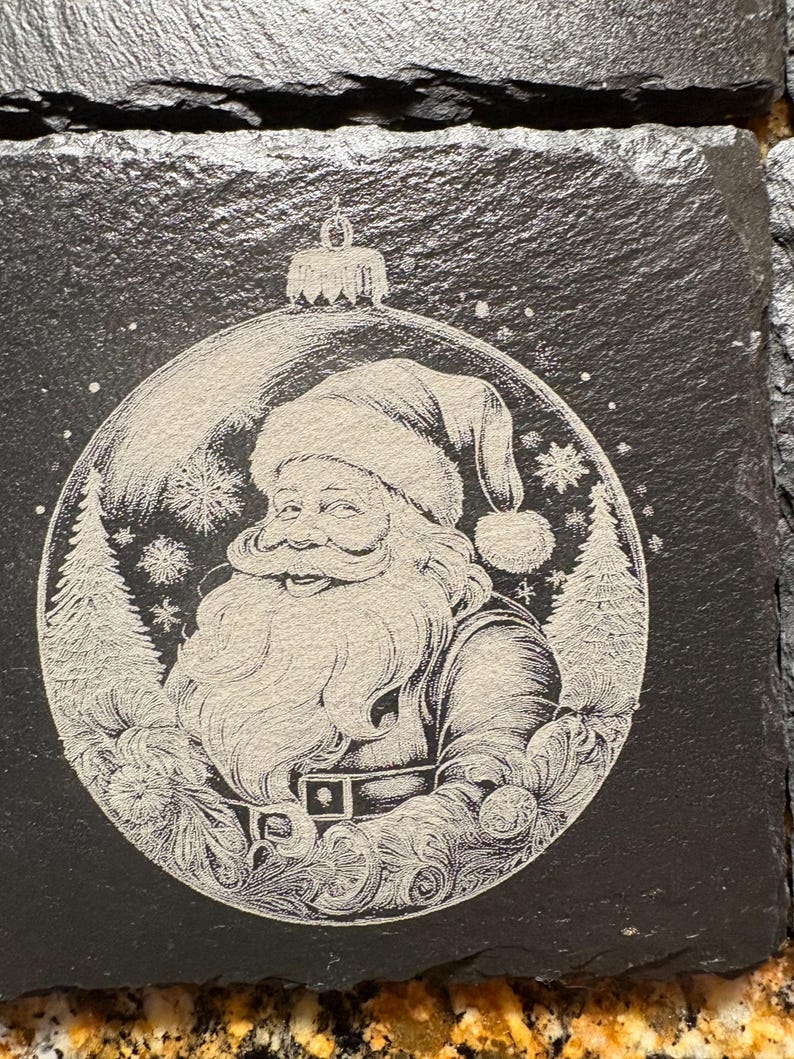 Pode incluir: Conjunto de bases quadradas de ard&oacute;sia preta com uma gravura detalhada do Pai Natal dentro de uma bola de Natal. O design inclui flocos de neve e pinheiros, criando um tema festivo de f&eacute;rias. As bases t&ecirc;m uma superf&iacute;cie natural e texturizada.