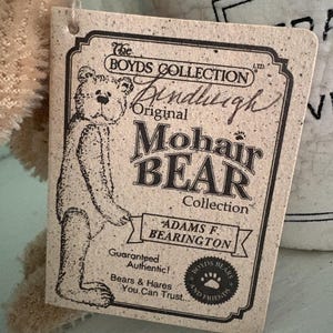 Pu&ograve; includere: Un'etichetta rettangolare beige di "The Boyds Collection" con uno schizzo di orso e le parole "Original Mohair Bear Collection". L'etichetta include anche il testo "Adams F. Bearington" e "Guaranteed Authentic!"