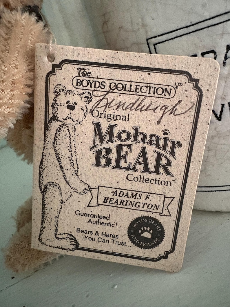 Pu&ograve; includere: Un'etichetta rettangolare beige di "The Boyds Collection" con uno schizzo di orso e le parole "Original Mohair Bear Collection". L'etichetta include anche il testo "Adams F. Bearington" e "Guaranteed Authentic!"