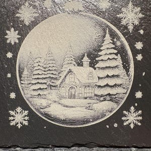 Pode incluir: Uma base de ard&oacute;sia preta com uma cena de inverno gravada em sua superf&iacute;cie. O design apresenta uma ilustra&ccedil;&atilde;o detalhada de uma casa de campo coberta de neve cercada por pinheiros, dentro de uma moldura circular, com flocos de neve espalhados.
