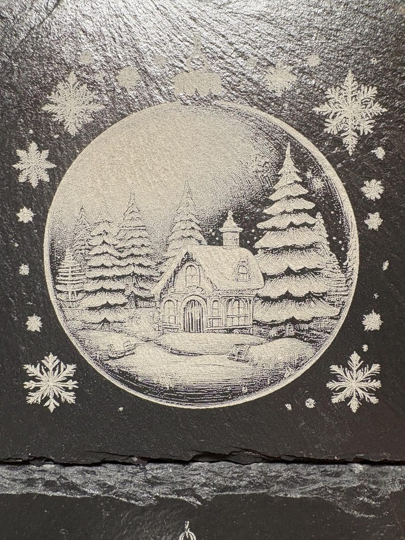 Pode incluir: Uma base de ard&oacute;sia preta com uma cena de inverno gravada em sua superf&iacute;cie. O design apresenta uma ilustra&ccedil;&atilde;o detalhada de uma casa de campo coberta de neve cercada por pinheiros, dentro de uma moldura circular, com flocos de neve espalhados.
