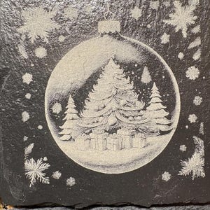 Pode incluir: Uma base de copo quadrada preta com um design gravado. O design apresenta uma &aacute;rvore de Natal e presentes dentro de uma bola transparente, rodeados por flocos de neve. A base de copo &eacute; feita de um material escuro e texturizado.