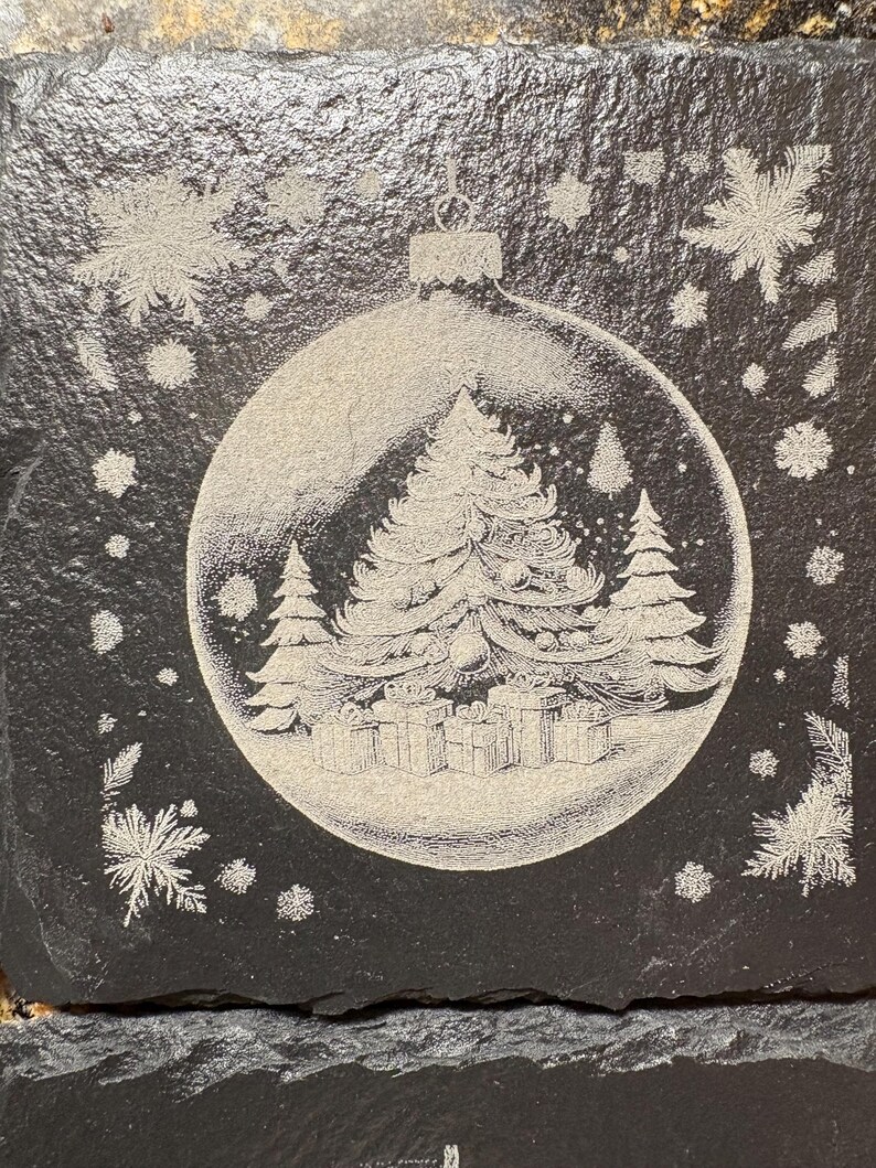 Pode incluir: Uma base de copo quadrada preta com um design gravado. O design apresenta uma &aacute;rvore de Natal e presentes dentro de uma bola transparente, rodeados por flocos de neve. A base de copo &eacute; feita de um material escuro e texturizado.