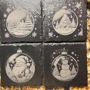 Pode incluir: Conjunto de quatro bases quadradas de ard&oacute;sia preta. Cada base apresenta uma cena de inverno gravada diferente dentro de uma forma de enfeite de Natal. As cenas incluem uma casa, &aacute;rvores, Papai Noel e um boneco de neve.