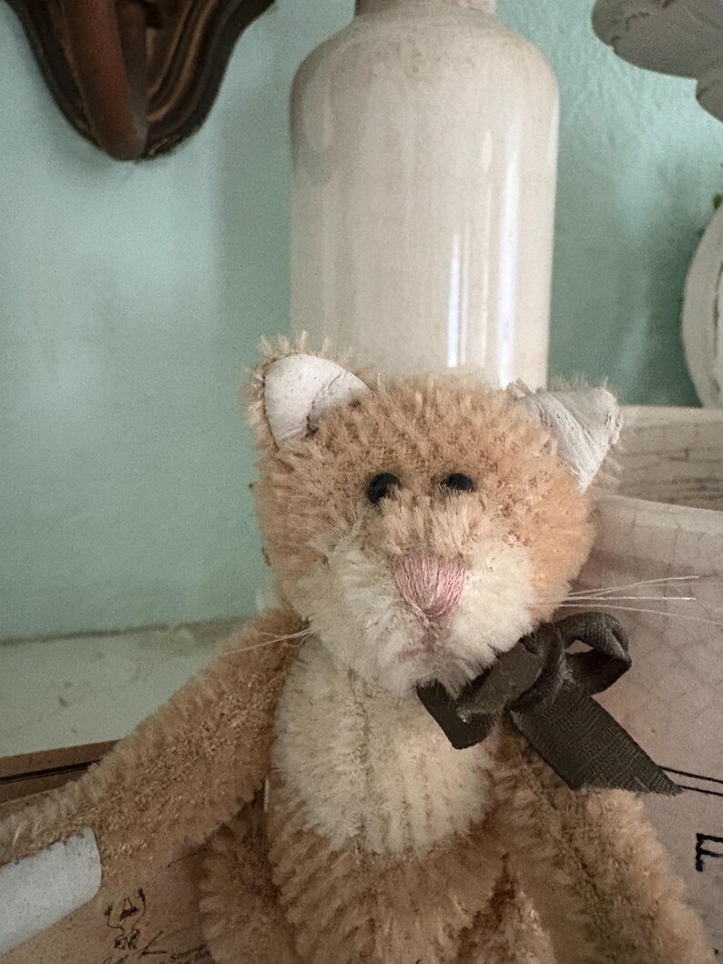 Pu&ograve; includere: Un giocattolo per gatti fatto a mano, in peluche, con pelo marrone chiaro e crema. Il gatto ha un naso rosa, occhi neri e un nastro marrone scuro legato a fiocco. Lo sfondo &egrave; morbido e sfocato.
