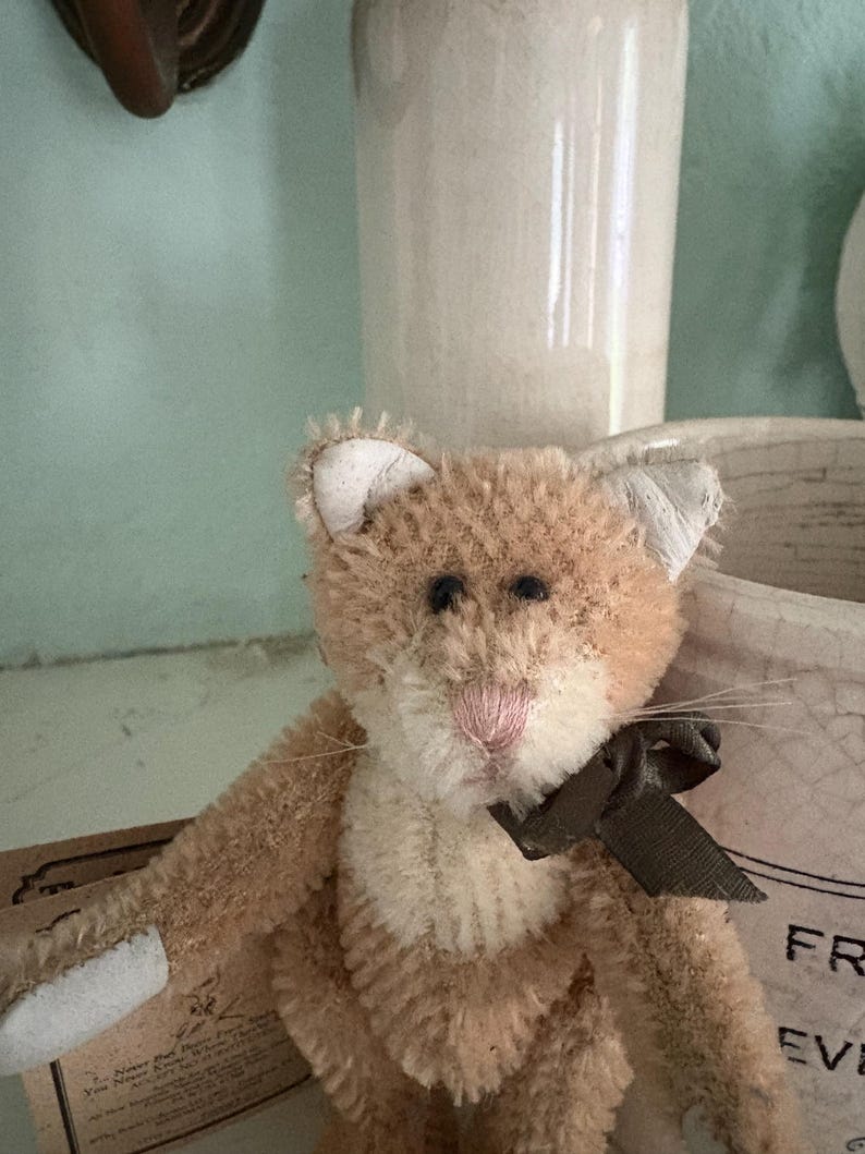Pu&ograve; includere: Un giocattolo a forma di gatto di peluche marrone chiaro con un fiocco marrone. Il gatto ha un naso rosa, occhi neri e orecchie interne bianche. Il giocattolo &egrave; realizzato in materiale morbido e strutturato.