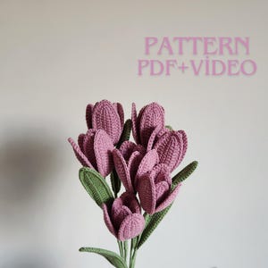 May include: A bouquet of crocheted tulips in shades of mauve and green. The handmade flowers have detailed petal and leaf textures. The image includes the text "PATTERN PDF+VIDEO" in pink.