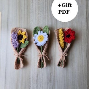 Puede incluir: Tres ramos de flores de ganchillo hechos a mano. Cada ramo presenta un diseño de flor diferente, incluyendo un girasol, una margarita y una amapola. Las flores están envueltas en papel de ganchillo color beige y atadas con una cuerda.