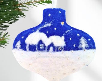May include: A teardrop-shaped felt Christmas ornament featuring a deep blue night sky with white embroidered stars and a snowy landscape with two small houses. The lower portion is white felt with scattered silver sequins and a white ribbon loop.