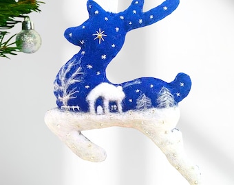 May include: A felted reindeer ornament in deep blue and white. The upper body features a starry night sky with a gold star, while the lower body depicts a snowy landscape with a small white house, trees, and a fence. It hangs from a white ribbon.