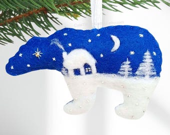 May include: A felt polar bear ornament featuring a deep blue night sky with a white crescent moon and stars. The bear's body is white with a snowy landscape, a small house, and evergreen trees stitched onto its back. It hangs from a white ribbon.