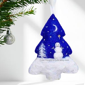 May include: A felt Christmas tree ornament in a deep blue color featuring a white snowman, a white pine tree, a crescent moon, and small stars. The base is covered in white felt with silver accents and the text CuteThingsForSoul.etsy.com.