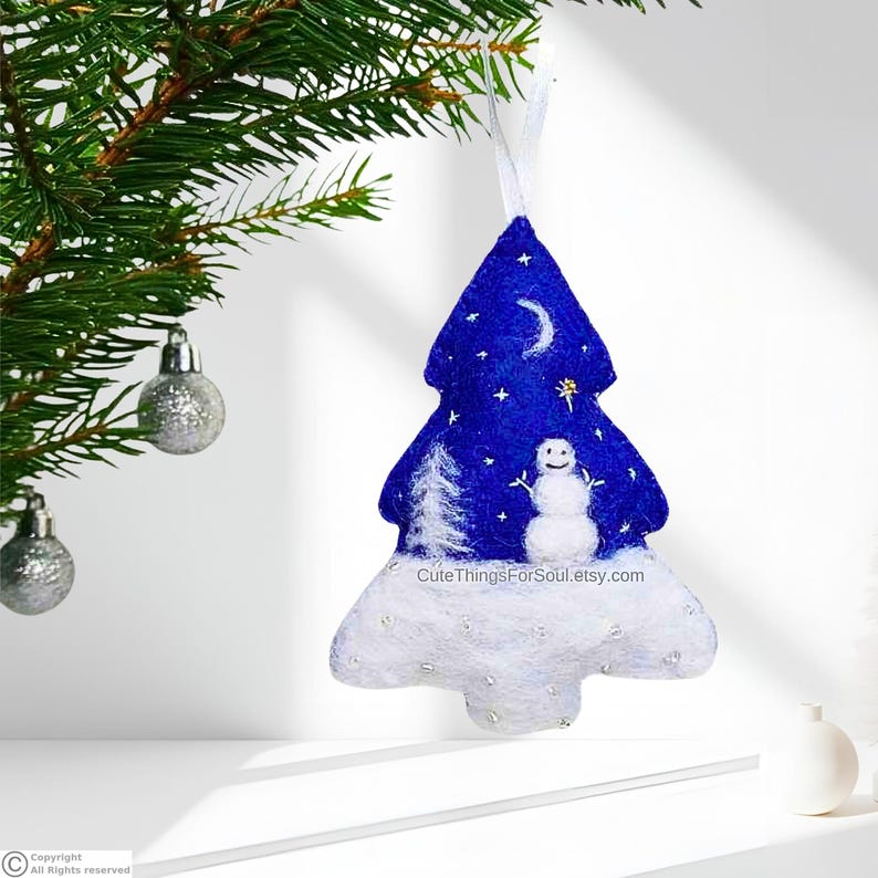 May include: A felt Christmas tree ornament in a deep blue color featuring a white snowman, a white pine tree, a crescent moon, and small stars. The base is covered in white felt with silver accents and the text CuteThingsForSoul.etsy.com.