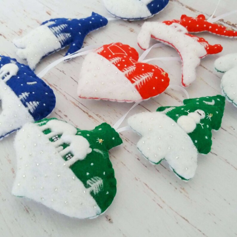 May include: A collection of felt ornaments in various shapes including hearts, trees, and animals. The ornaments feature red, blue, green, and white colors with embroidered details, white stitching, and small white beads attached to white ribbons.