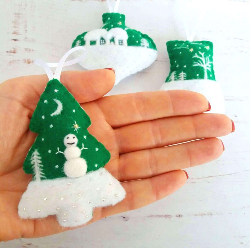 May include: Three felt Christmas tree ornaments in green and white. The foreground ornament is shaped like a tree with a white snowman, a crescent moon, and stars. The other two ornaments feature a house and a forest scene, all with white ribbon loops.
